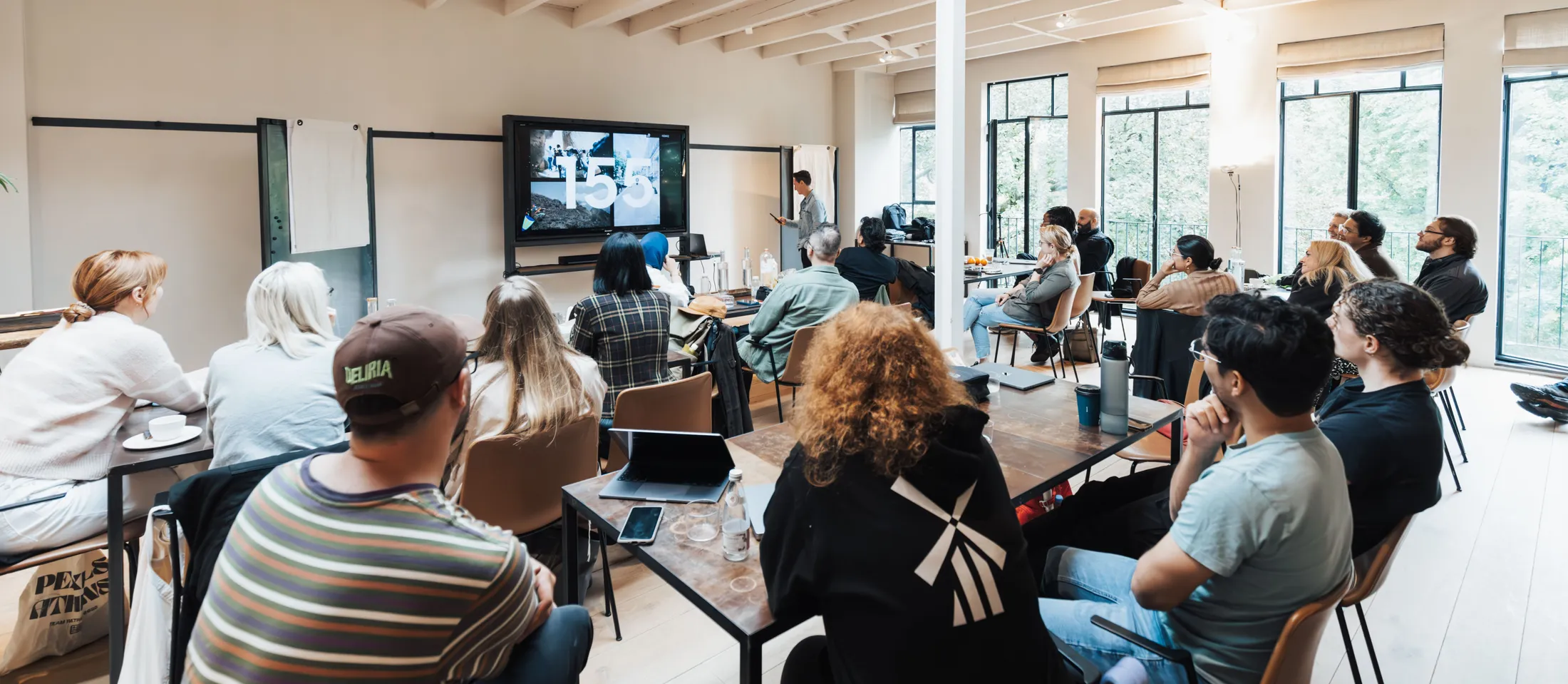 A room of professionals seated at tables in a bright loft studio, turned toward a presenter and a wall-mounted screen.