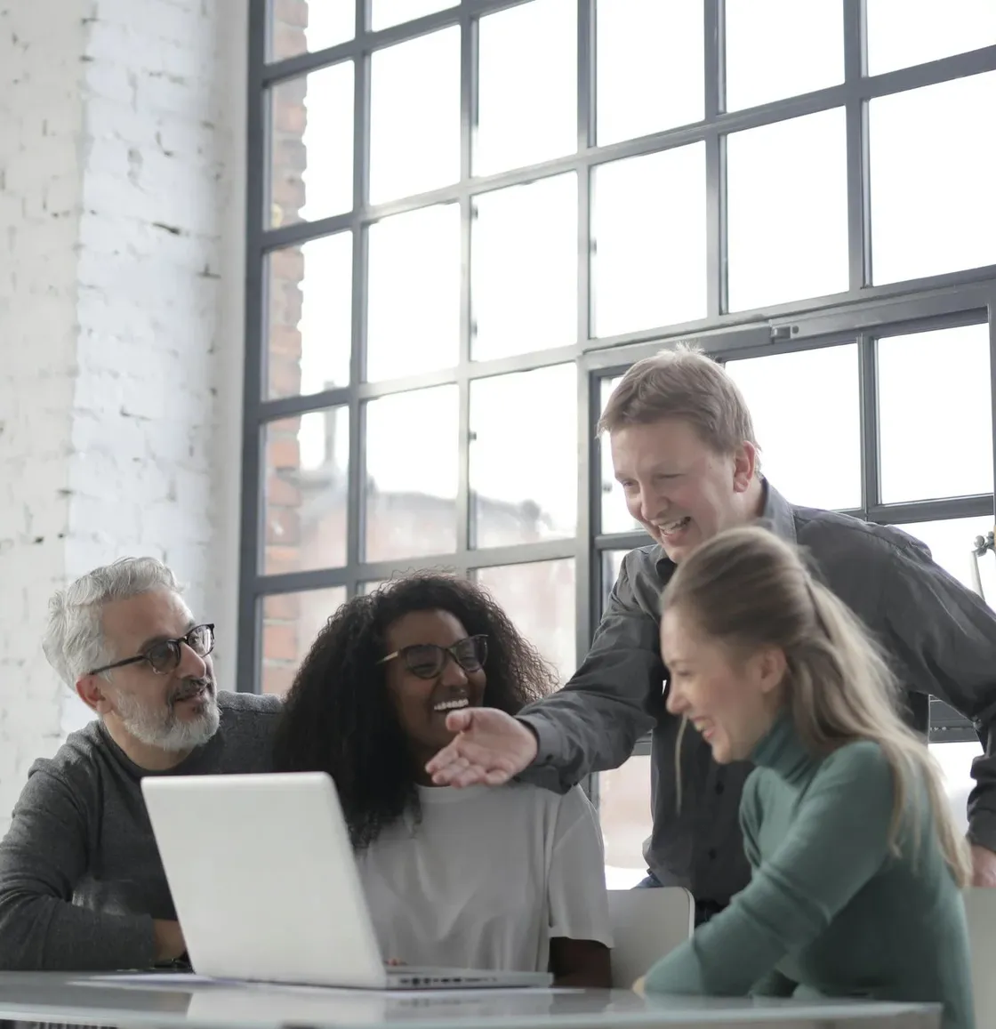 Four colleagues gathered around a laptop in a bright loft office, laughing as one of them reaches across the table.